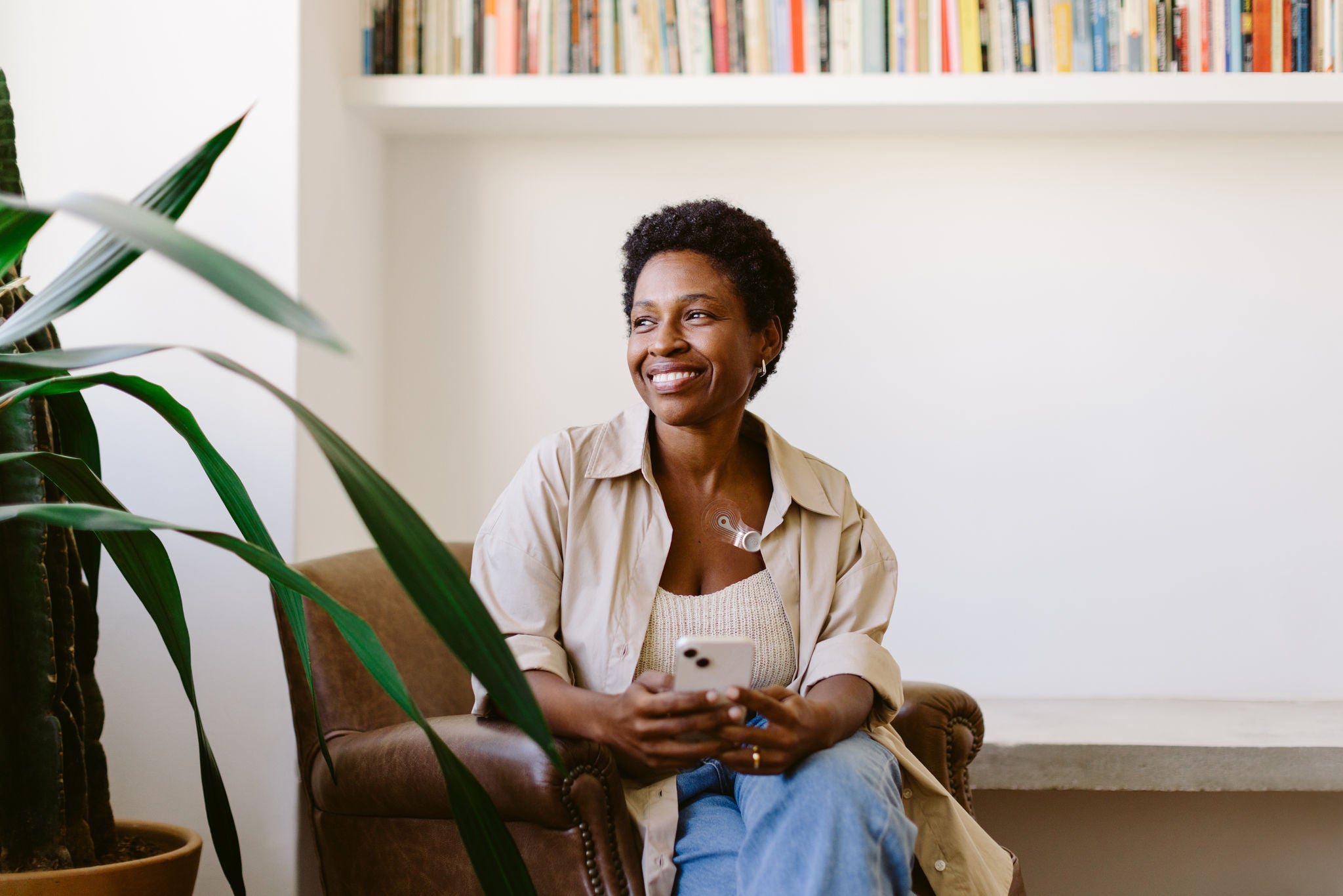 Afro haired black woman sitting on a couch, surrounded by a bookshelf. She holds a mobile phone, looking thoughtful. Mature black woman enjoying her leisure time at home.