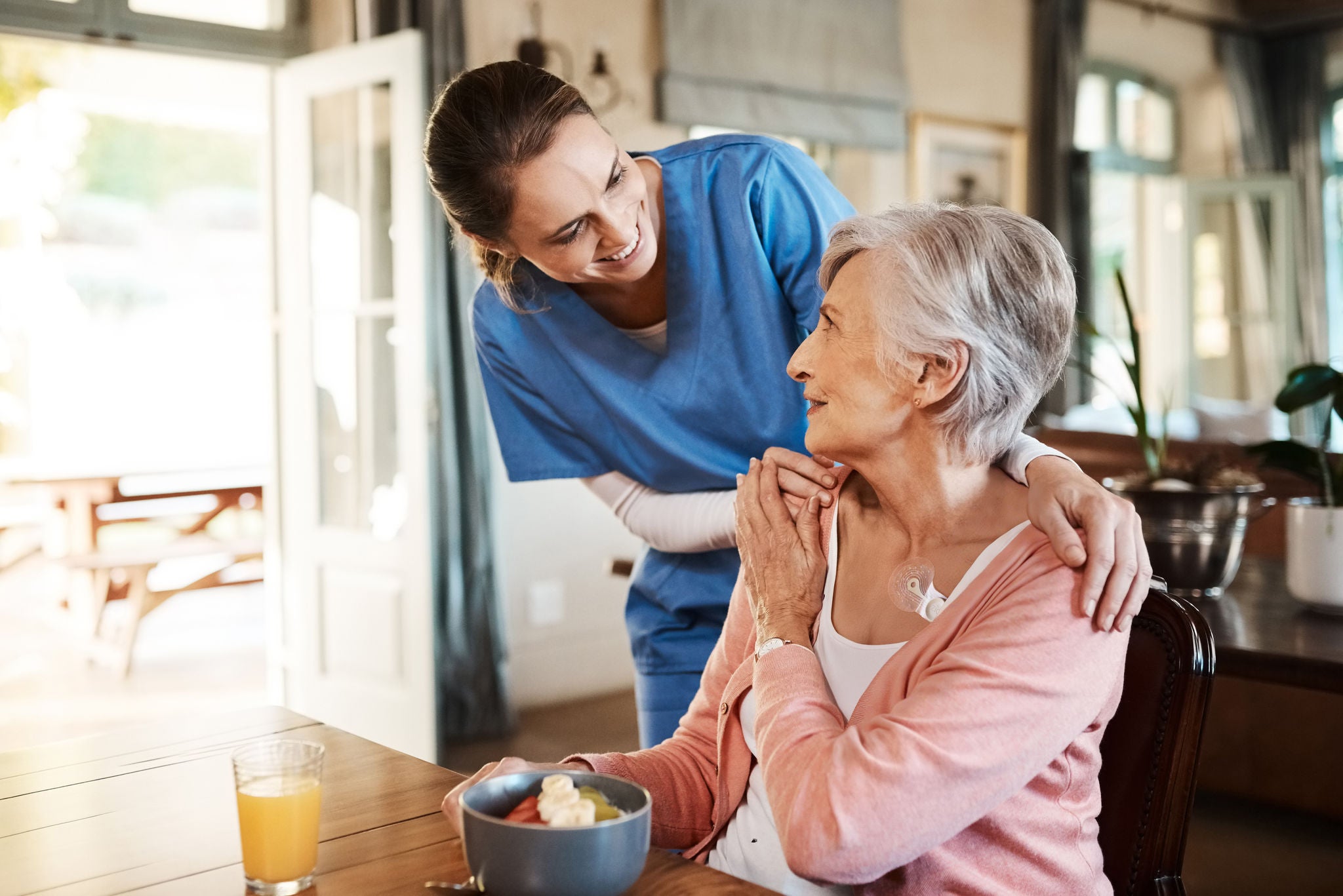 Healthcare, elderly woman with nurse with breakfast at her home and at the table in living room. Support or communication, caregiver and conversation with medical person with senior or old female